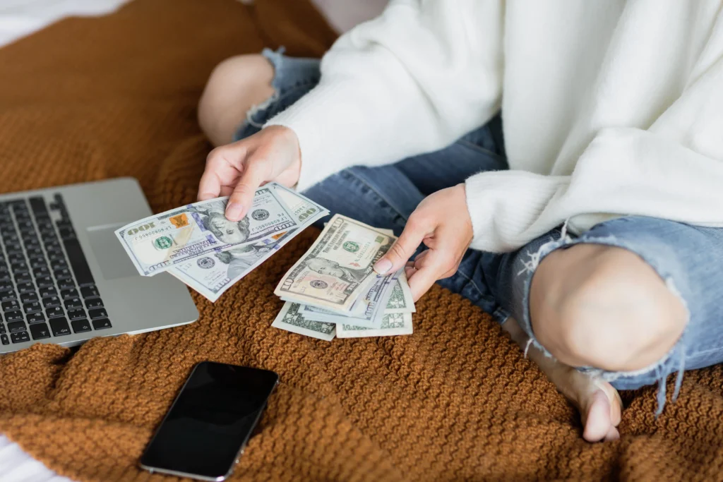 Persona sentada en casa con laptop y celular mientras cuenta billetes de dólar